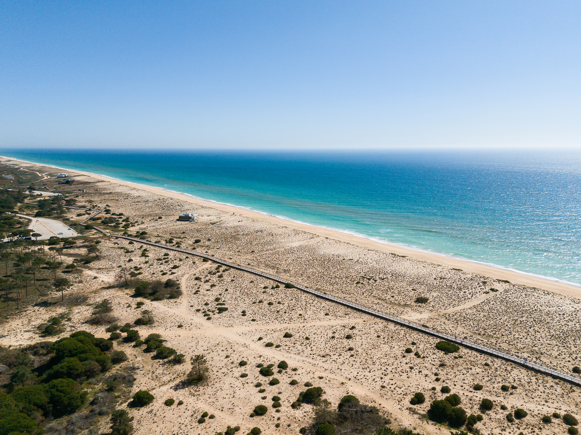 Aerial view of a wide Algarve beach with golden dunes, a wooden boardwalk, and calm Atlantic waters under a clear blue sky.